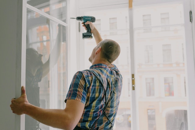 contractor in a blue plaid shirt using a drill to fix a window hinge